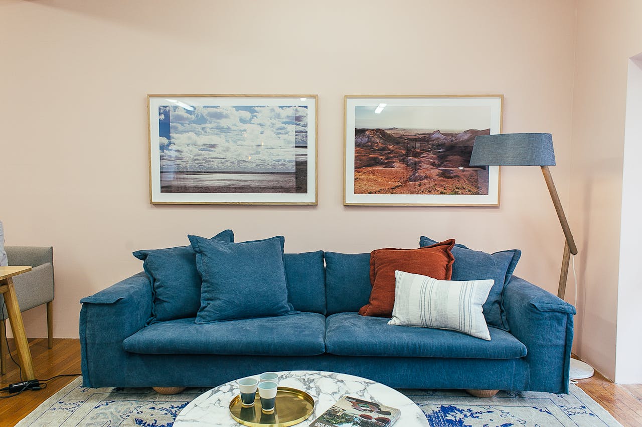 Big comfortable blue sofa with soft cushions placed in modern living room with round marble table near wall decorated with framed pictures